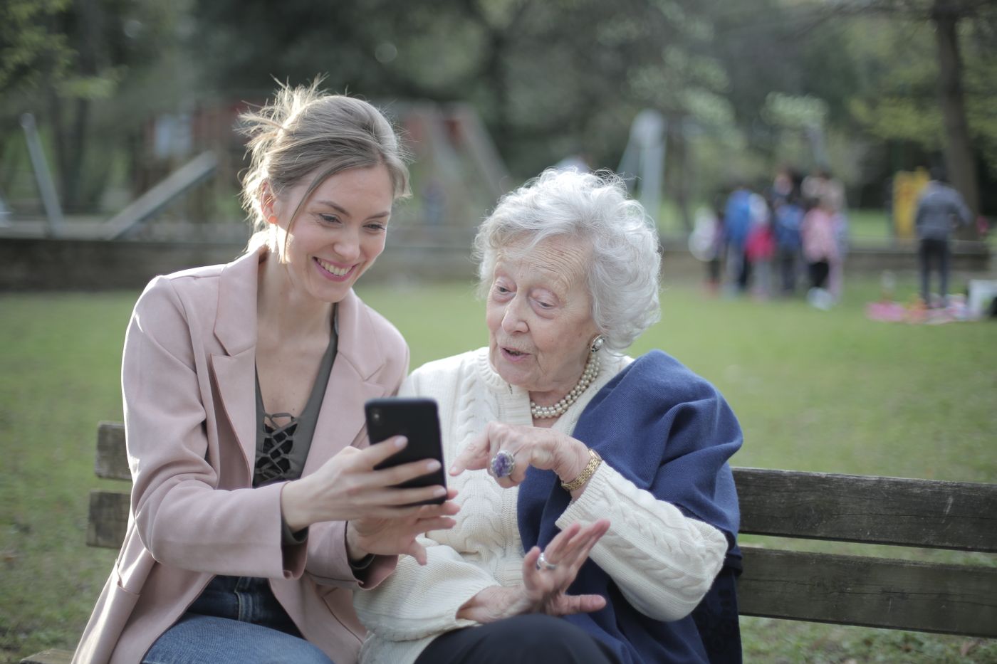A younger woman and an older woman sitting on a park bench, looking at a phone screen together